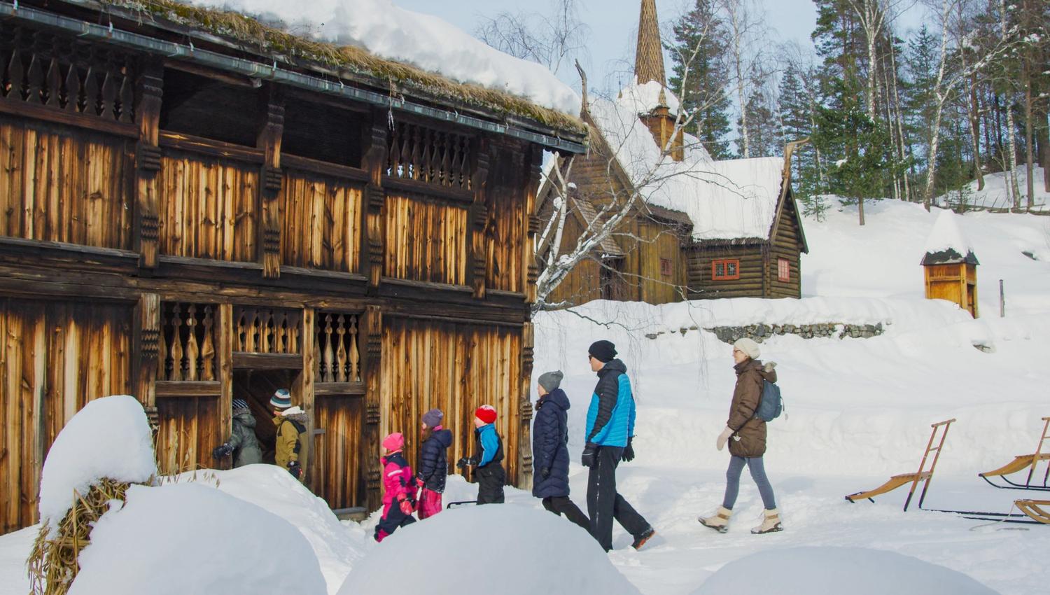 Voksne og barn går inn i tømmerhus ved Garmo stavkirke.