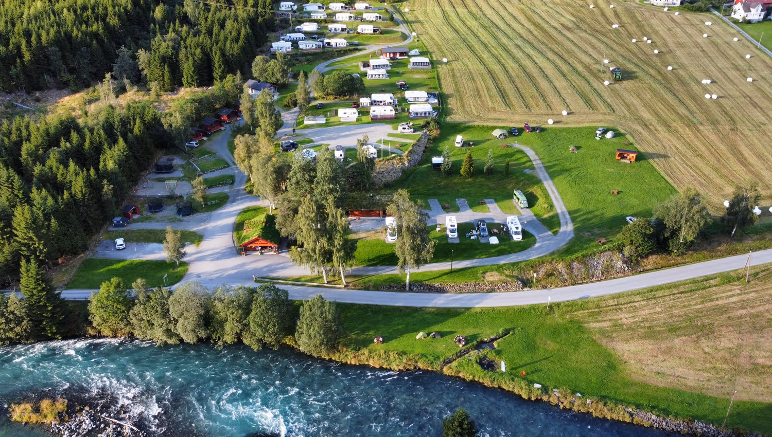 Luftfoto av ein campingplass med bubilar og telt ved ei elv, veg, skogkant og slåtteåkrar med rundballar..