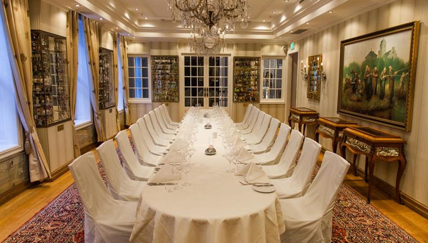 Long table set for dining with white tablecloth, chairs, paintings on the walls, chandelier in the ceiling
