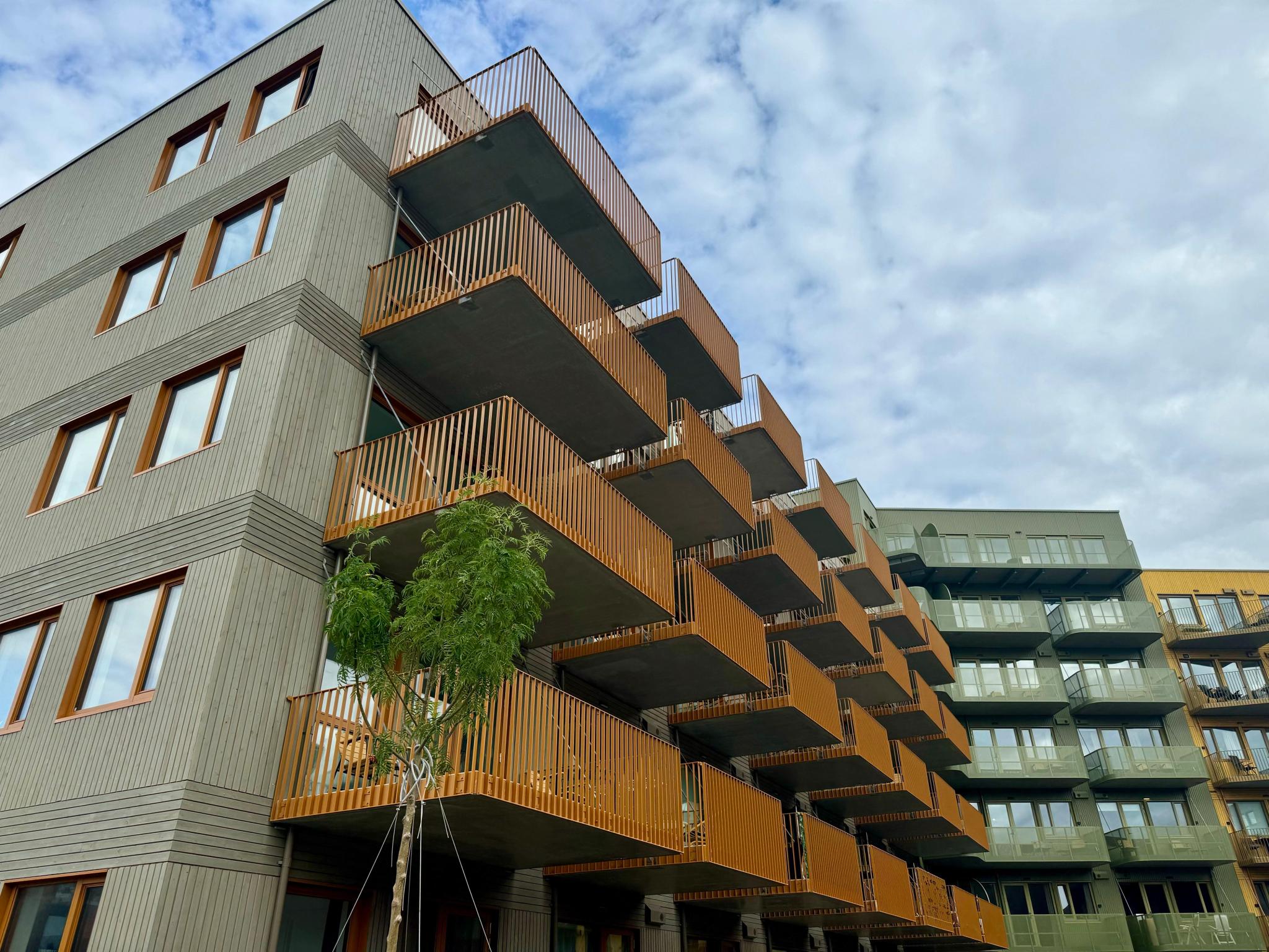 Moderne leilighetsbygg med grå fasade og trepanel på balkonger under delvis skyet himmel.