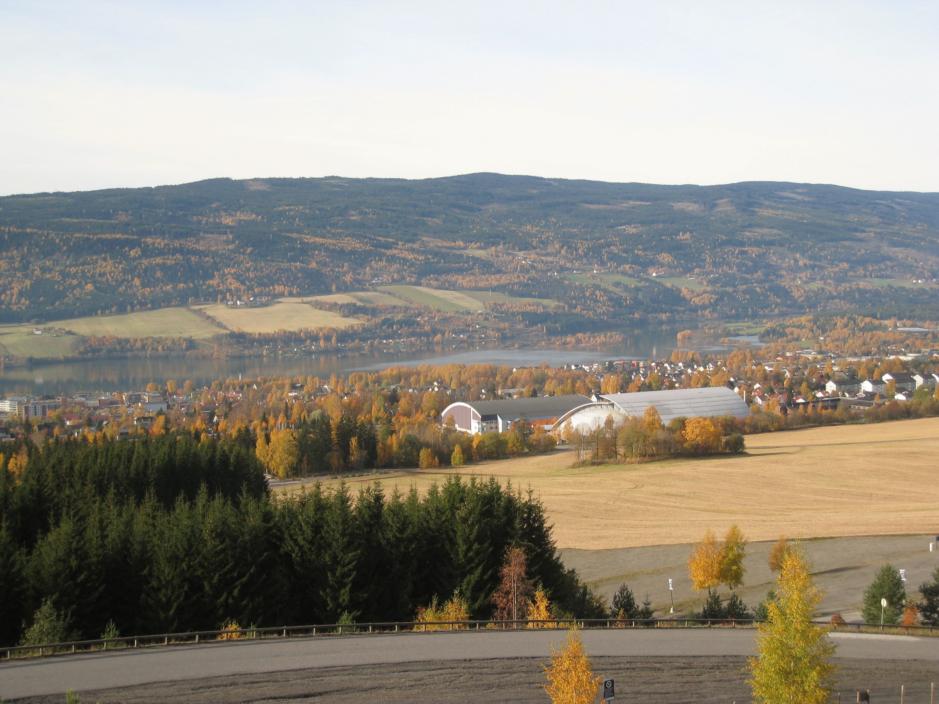 Panorama av åkre, skog og et tettsted ved en innsjø, med åser i bakgrunnen og høstfarger i trærne..