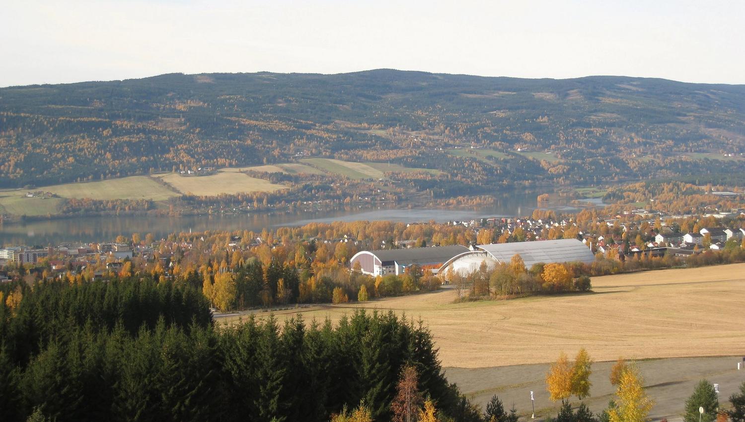 Panorama av åkre, skog og et tettsted ved en innsjø, med åser i bakgrunnen og høstfarger i trærne..