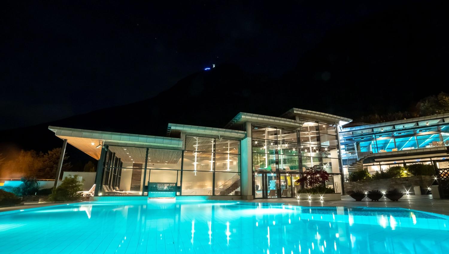 Illuminated outdoor swimming pool in front of a modern glass building at night, with hills silhouetted in the background..