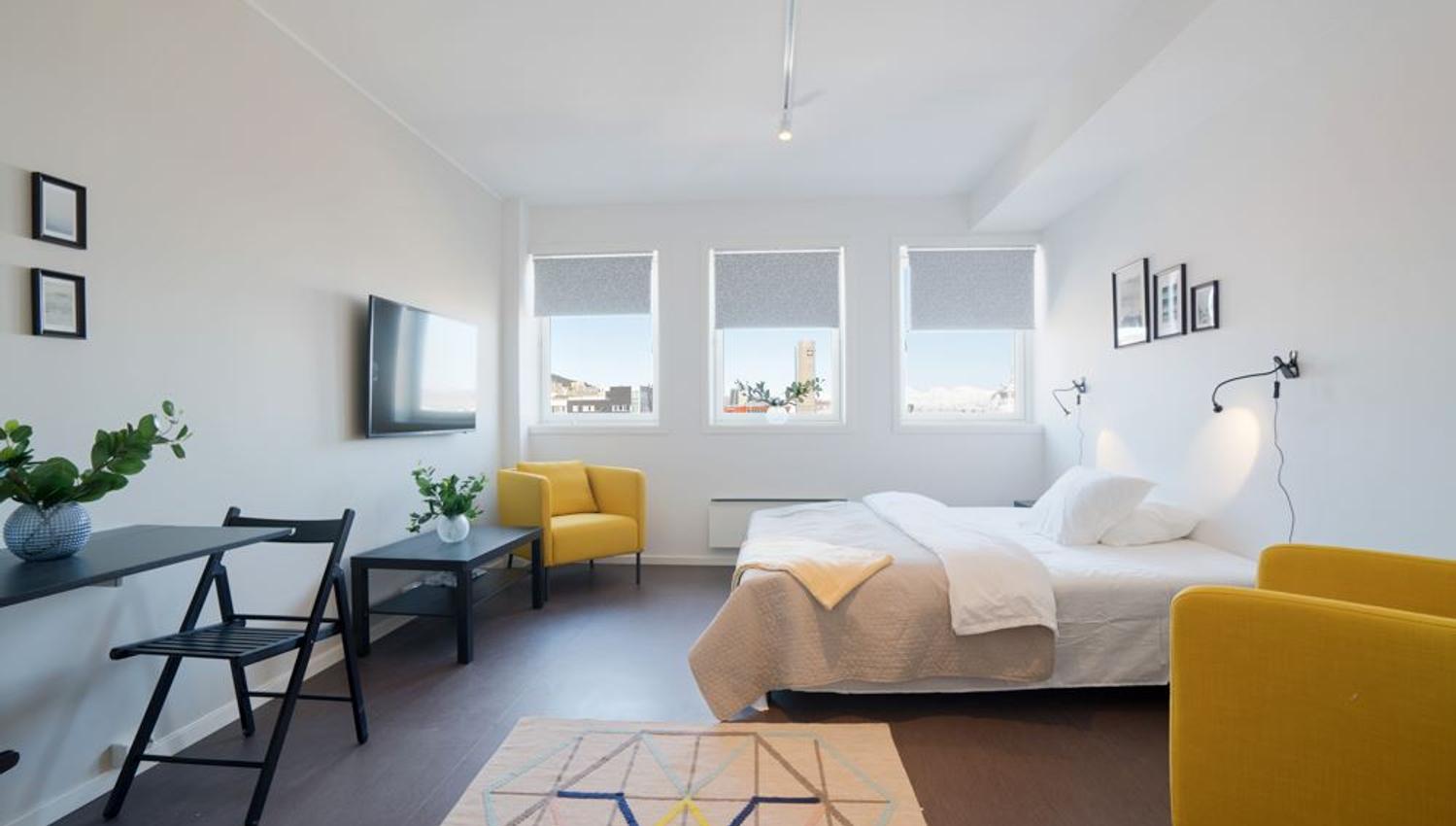 Lyst soverom med seng, skrivebord, lenestoler og store vinduer.