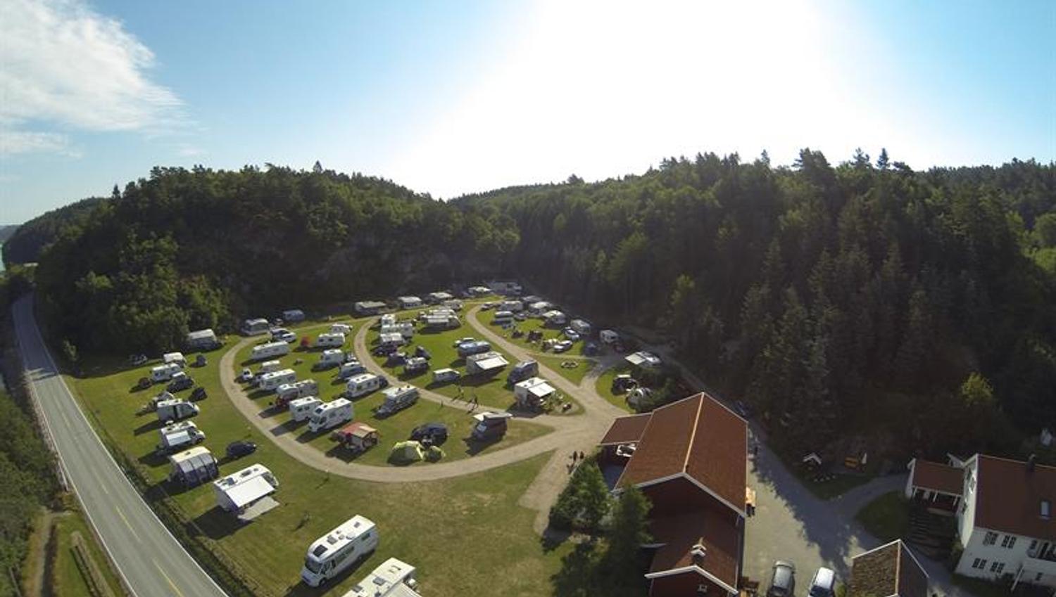 Drohnenaufnahme des Campingplatzes mit Wohnmobilen/Wohnwagen in Reihen, mit Wegen zwischen den Reihen, einer Hauptstraße in der Nähe und Wald im Hintergrund.