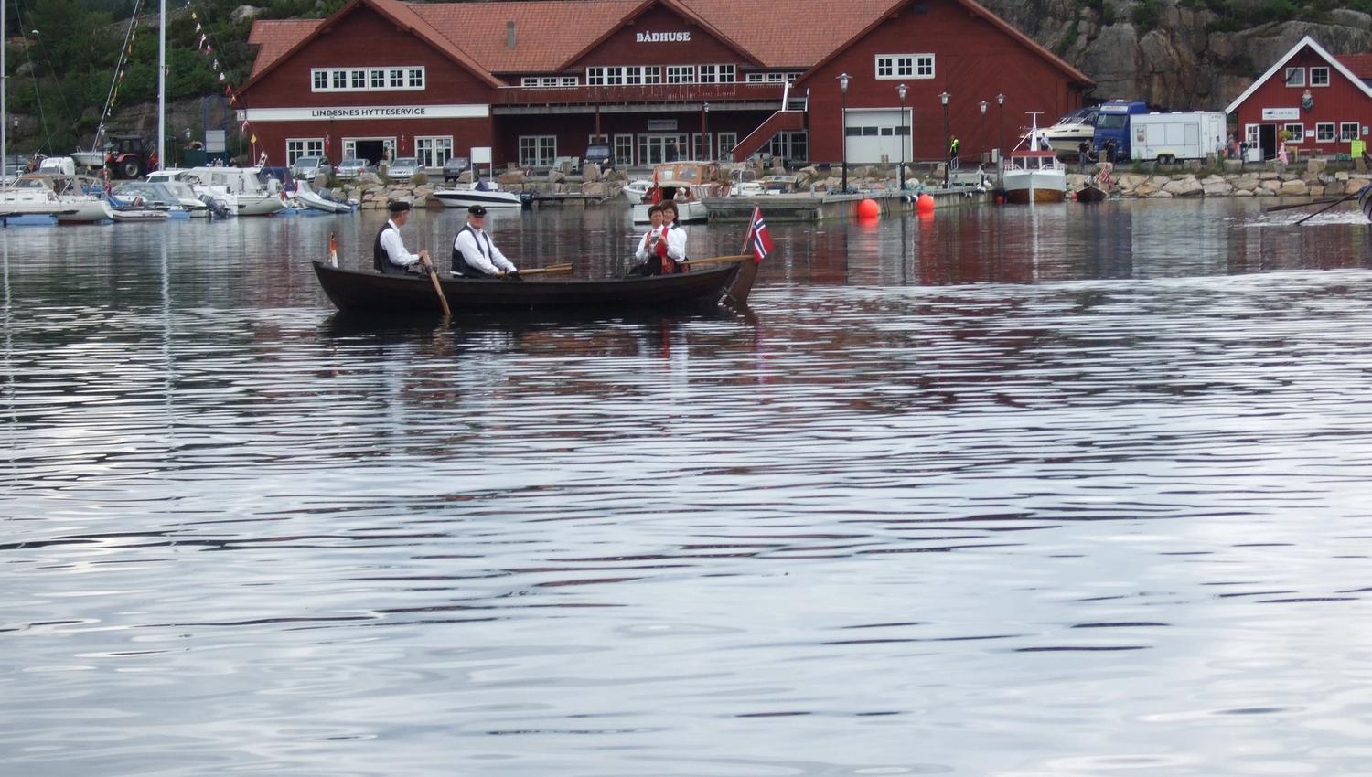 Small wooden boat on the water with 2 men and 2 women in traditional Norwegian costumes (bunads), and the Norwegian flag at the tip of the boat, in fr
