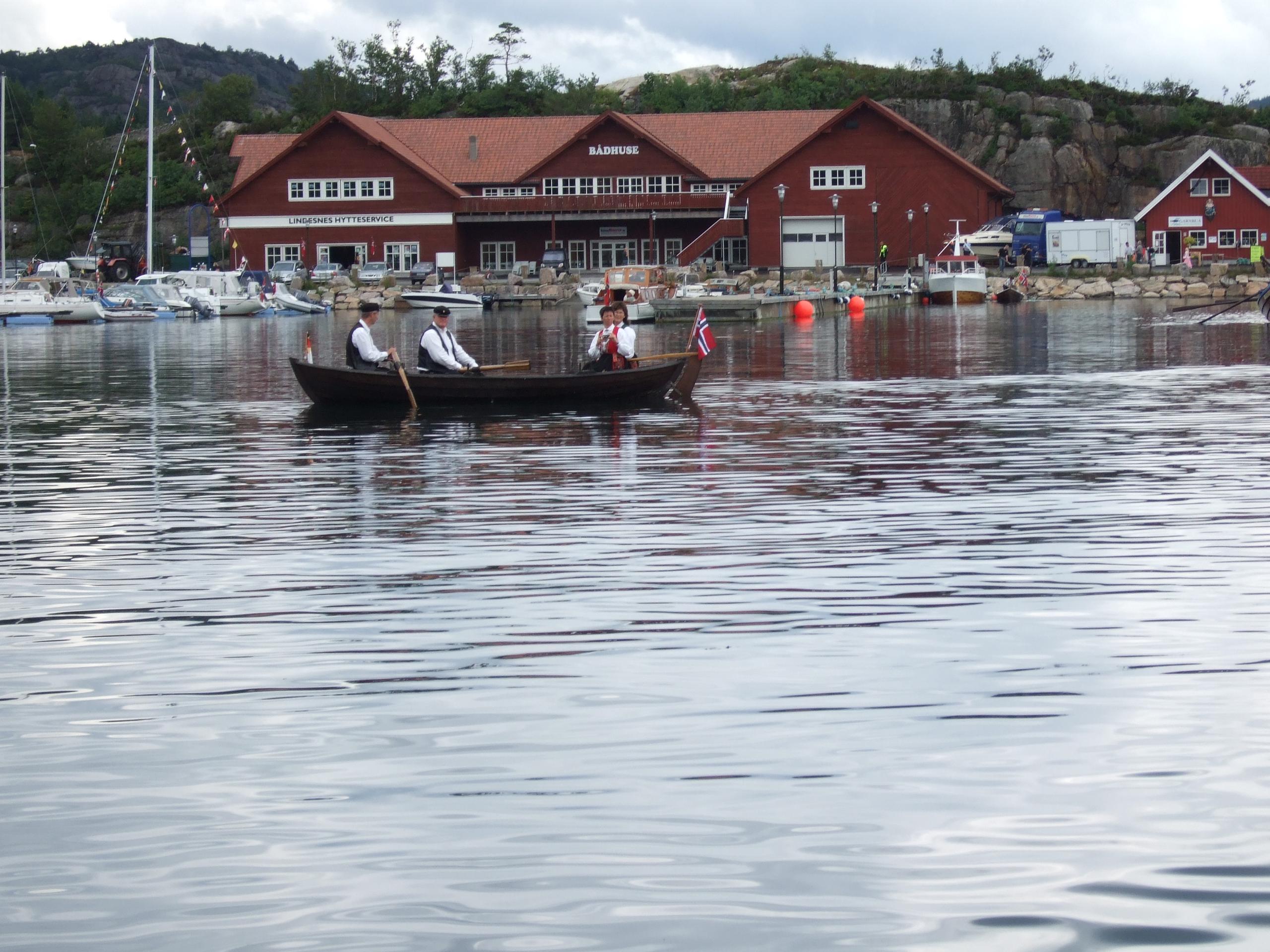 Small wooden boat on the water with 2 men and 2 women in traditional Norwegian costumes (bunads), and the Norwegian flag at the tip of the boat, in fr