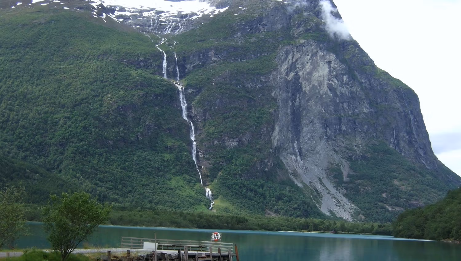 Ramnefjellsfossen waterfall runs down a green mountain with patches of snow, above a calm lake and a small wooden dock..