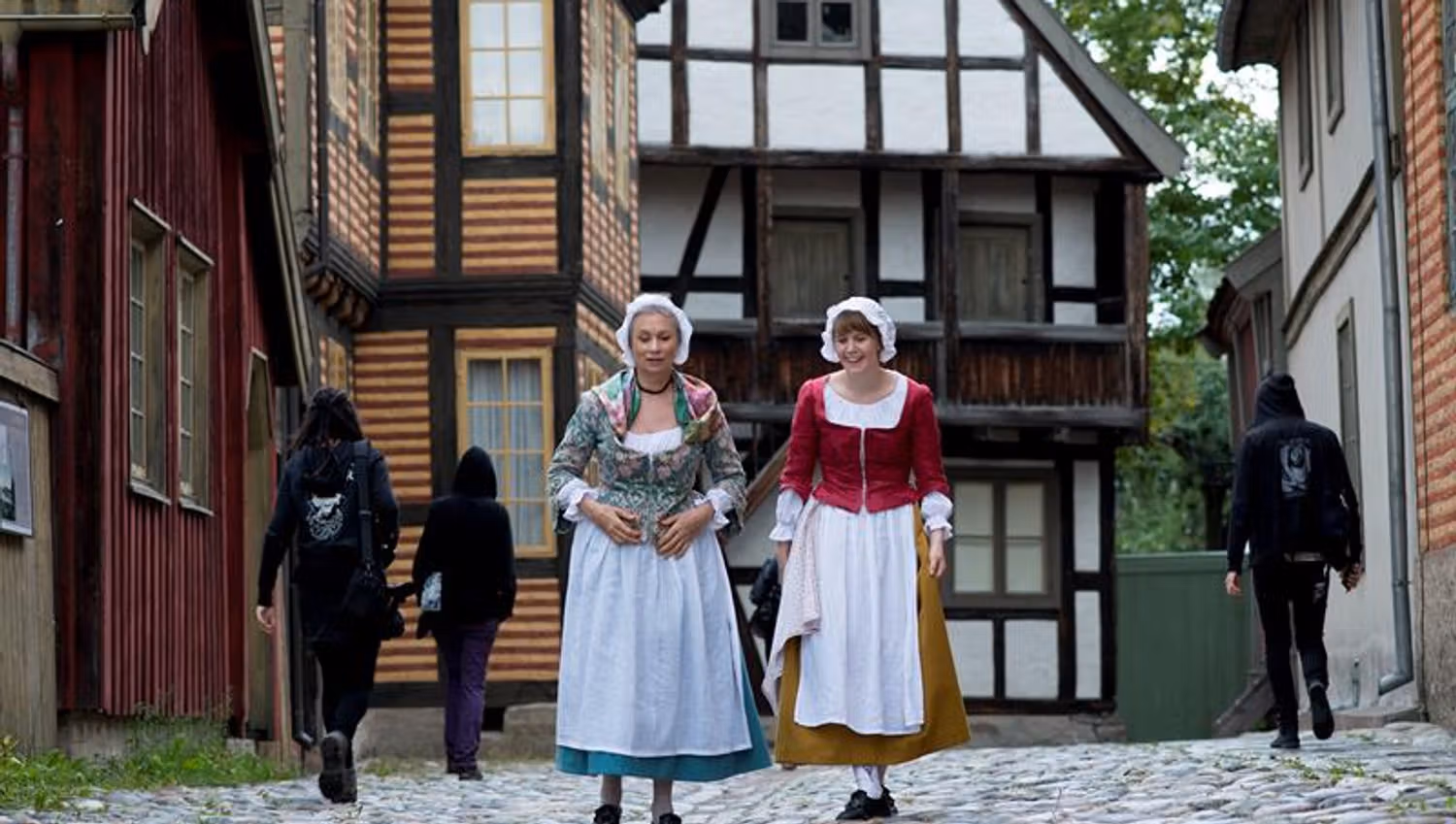 Two women in traditional clothing walking by old houses. Norwegian Museum of Cultural History