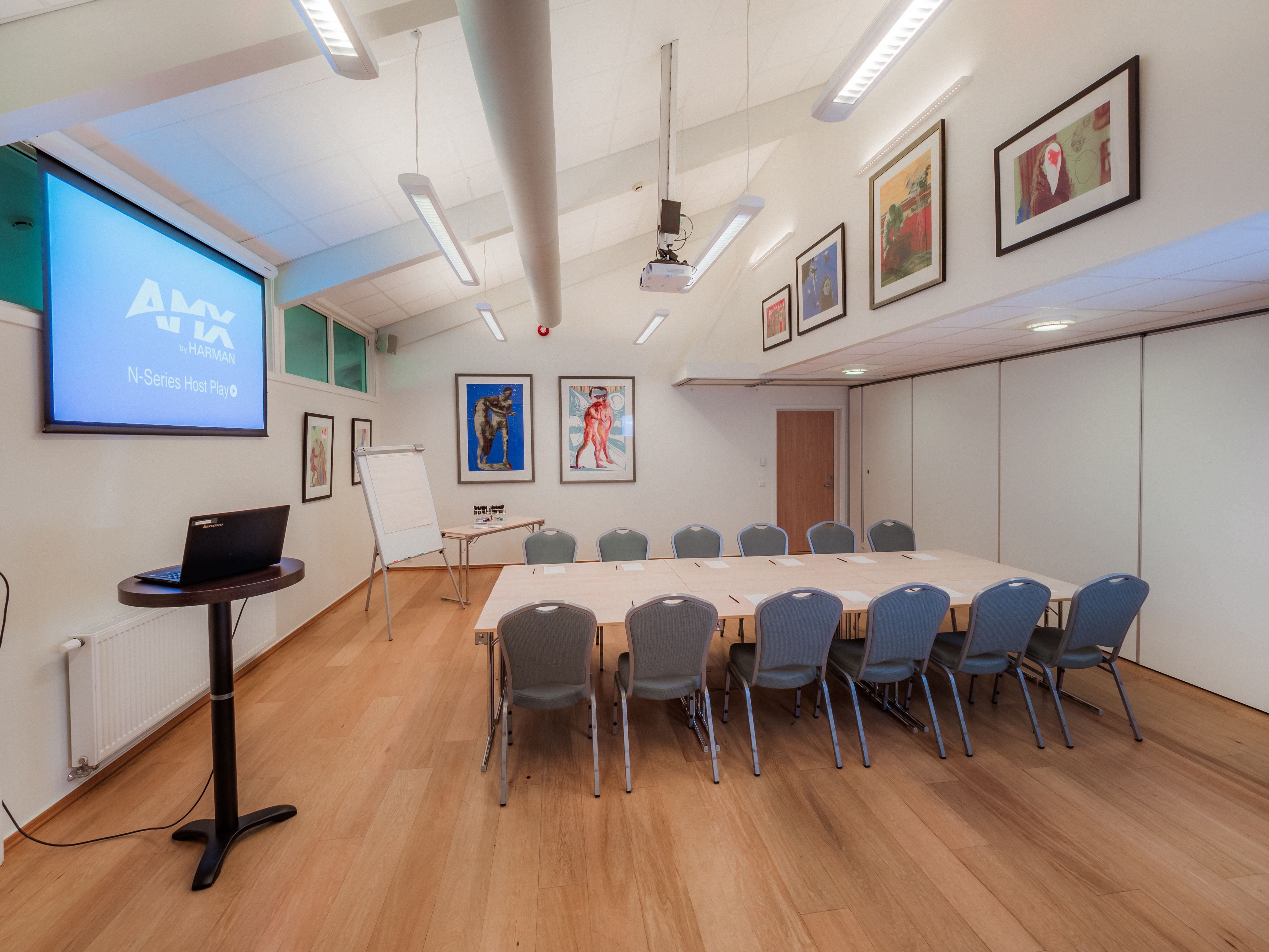 Modern meeting room with U-shaped tables and chairs, a projector screen, flip chart, and framed wall art.