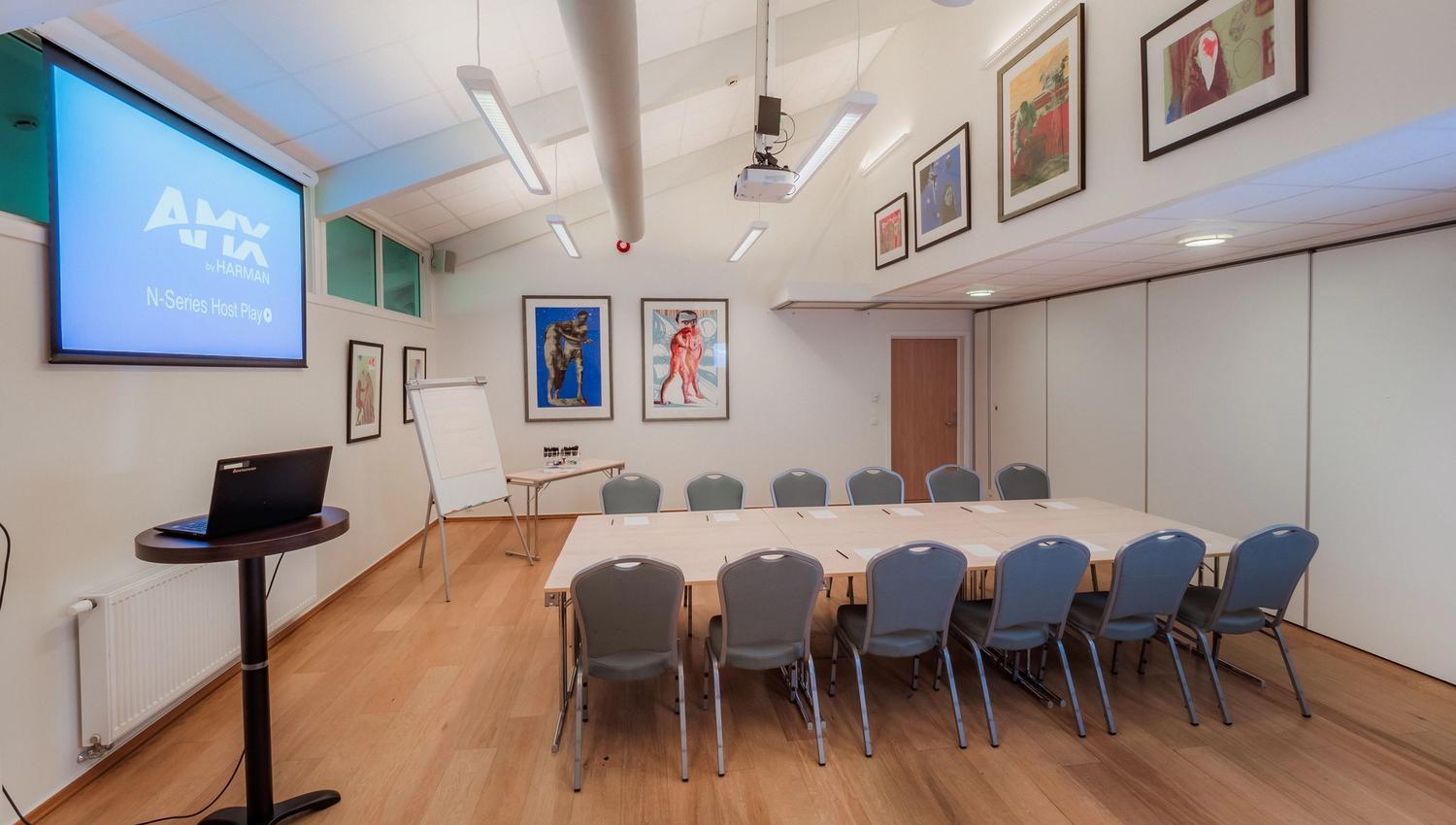 Modern meeting room with U-shaped tables and chairs, a projector screen, flip chart, and framed wall art.