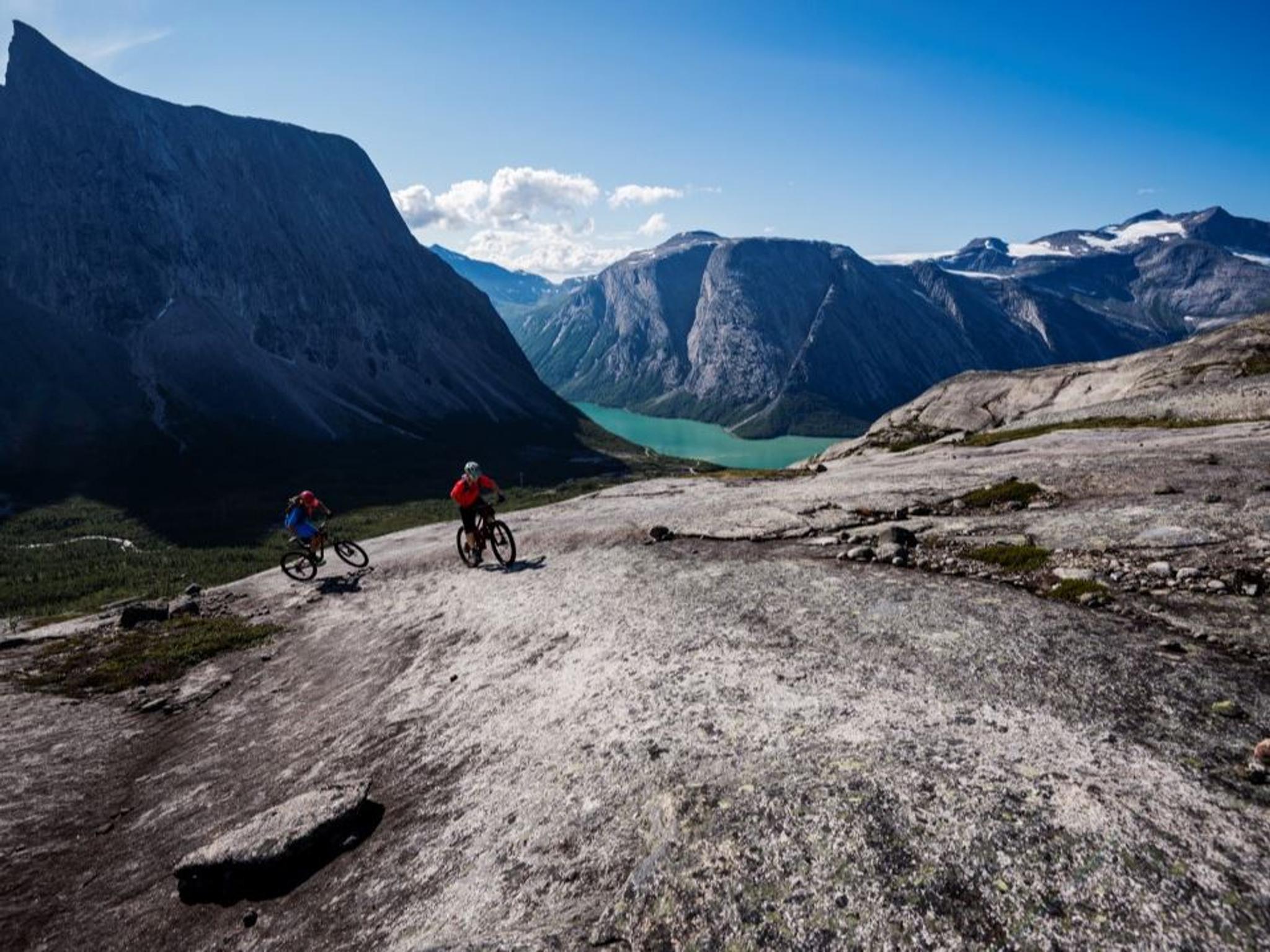 Two mountain bikers ride over bare mountain slabs with a turquoise lake and steep mountains in the background.