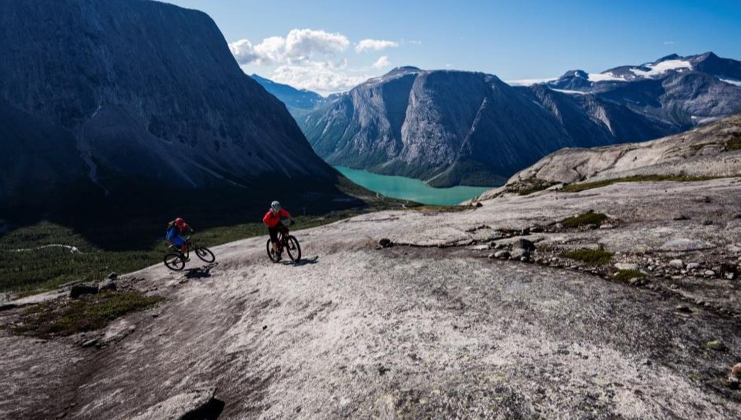 Two mountain bikers ride over bare mountain slabs with a turquoise lake and steep mountains in the background.