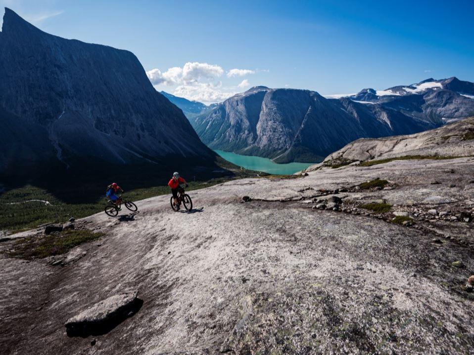 Two mountain bikers ride over bare mountain slabs with a turquoise lake and steep mountains in the background.