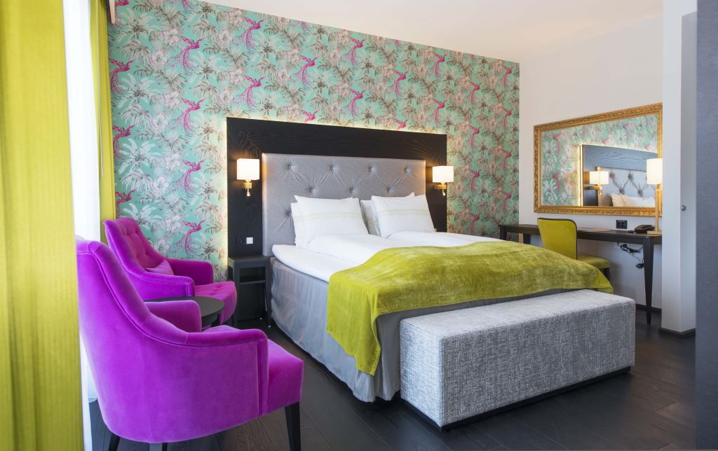 Interior of hotel room: Double bed, purple armchairs, floral wallpaper.
