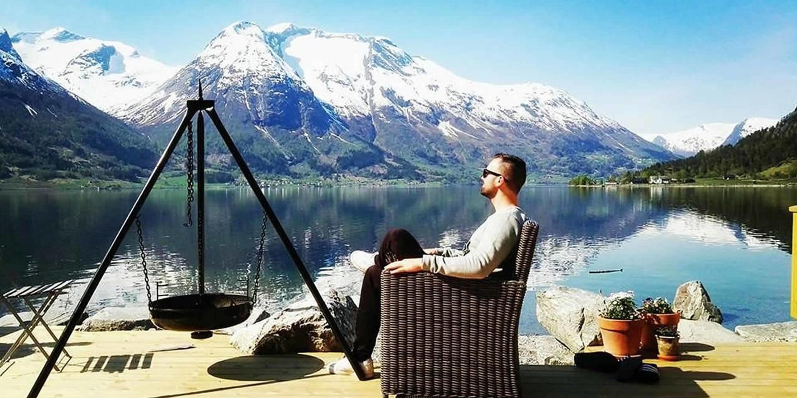 Ein person sit i ein flettstol på ei brygge på Vollsnes Feriehus ved stille fjordvatn med snødekte fjell i bakgrunnen..