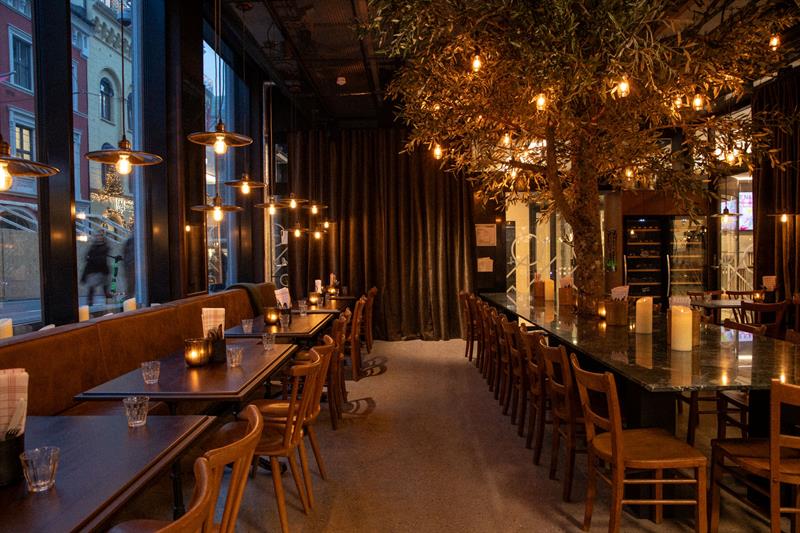 En restaurant med moderne glassfasade og et varmt, stemningsfullt interiør med trebord, hengende lamper og dekkede bord langs vinduene.