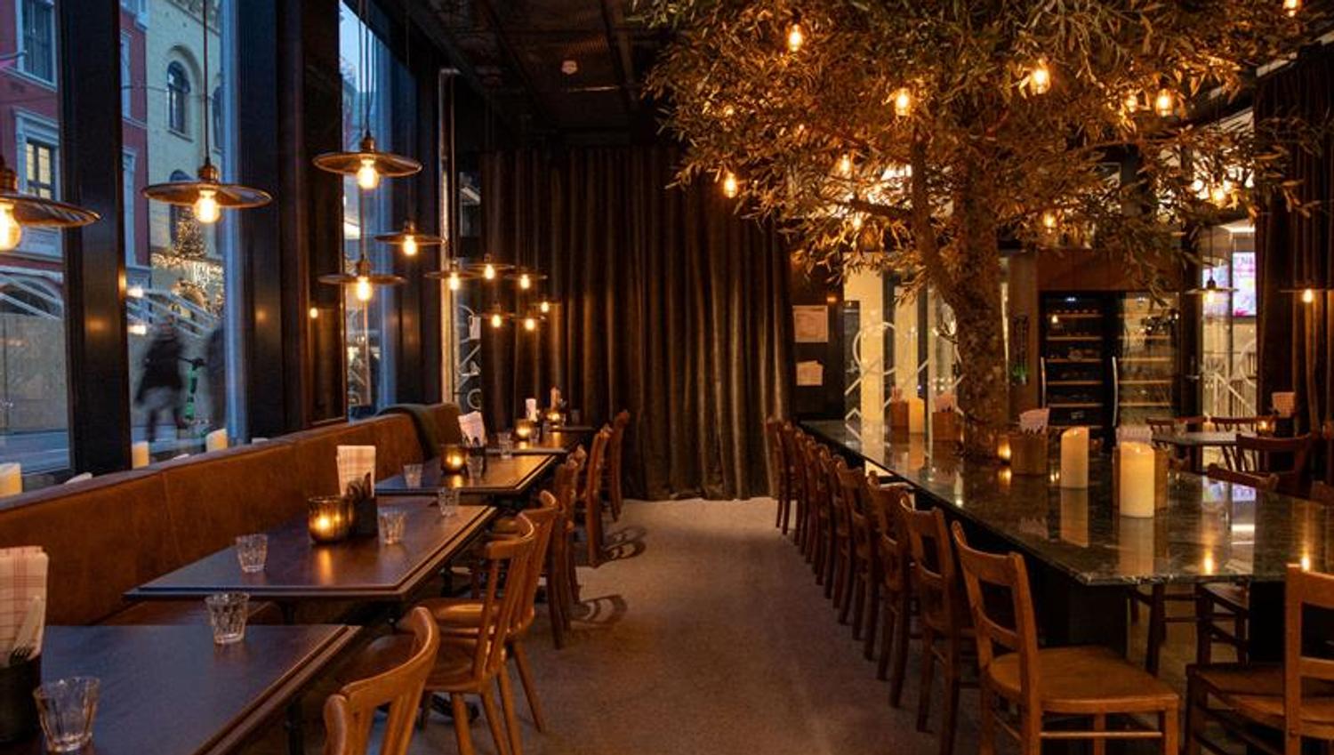 En restaurant med moderne glassfasade og et varmt, stemningsfullt interiør med trebord, hengende lamper og dekkede bord langs vinduene.