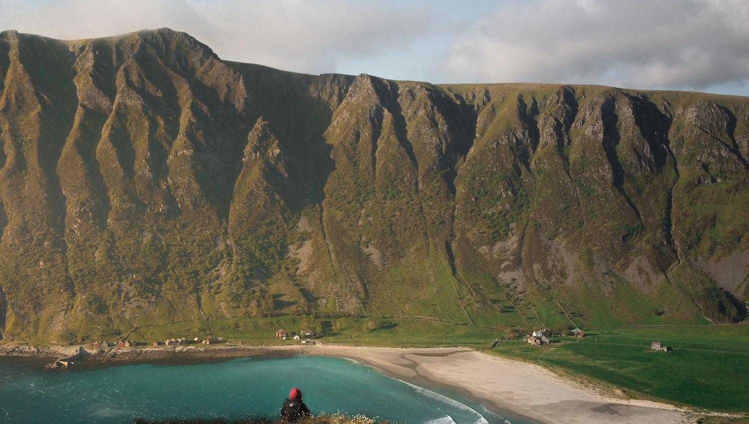 Ein person sit på ein kolle og ser ned mot turkis hav, sandstrand, grøne marker og bratte fjell under skyer..