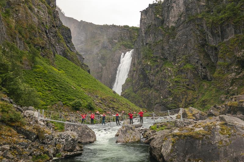 People in red clothing, waving norwegian flags, standing on the new bridge with the waterfall seen in the background.
