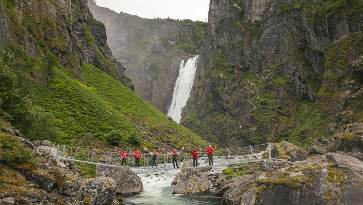 People in red clothing, waving norwegian flags, standing on the new bridge with the waterfall seen in the background.