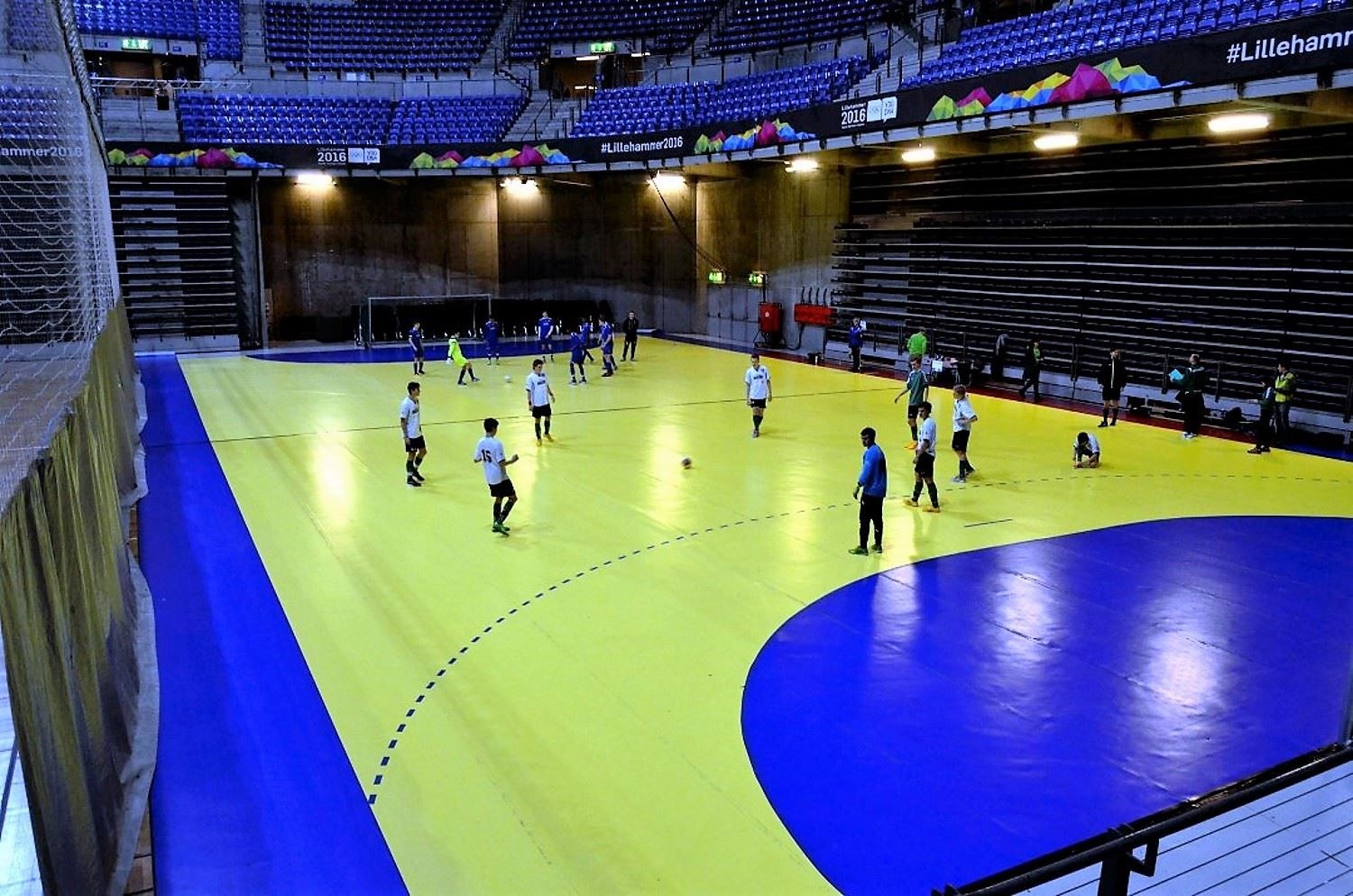 Innendørs håndballbane med gult og blått gulv, flere spillere som varmer opp, og tomme tribuner i bakgrunnen..