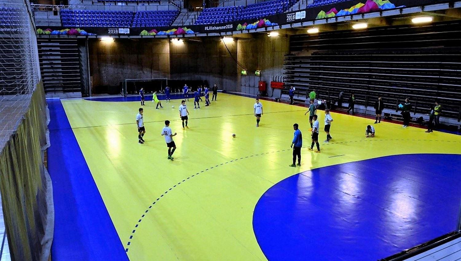 Innendørs håndballbane med gult og blått gulv, flere spillere som varmer opp, og tomme tribuner i bakgrunnen..