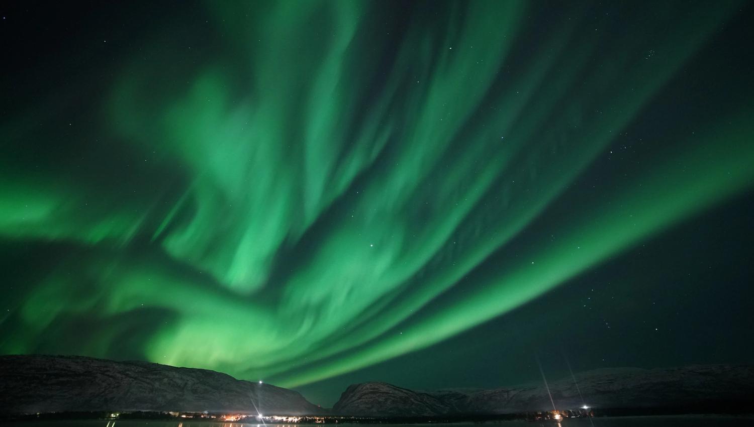 Witness the breathtaking display of the northern lights as they dance across the starry sky, casting a mesmerizing green glow over the tranquil waters. This stunning view captures the magic of nature in a remote, peaceful setting.