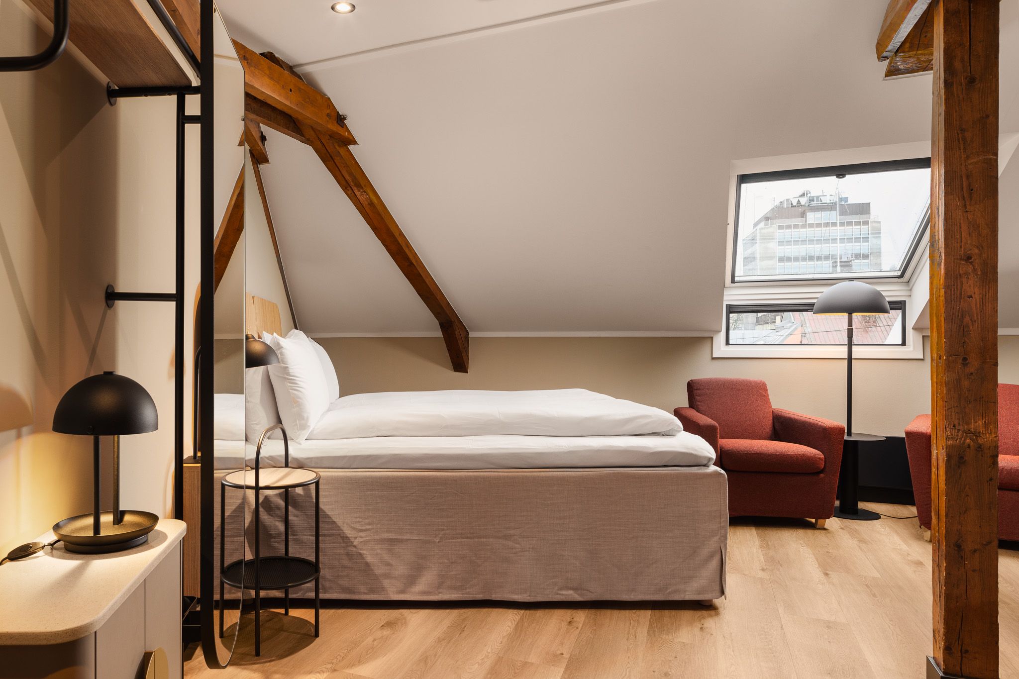 A hotel room interior with a double bed, a window, brown armchairs and a brown ceiling beam running along the curved ceiling