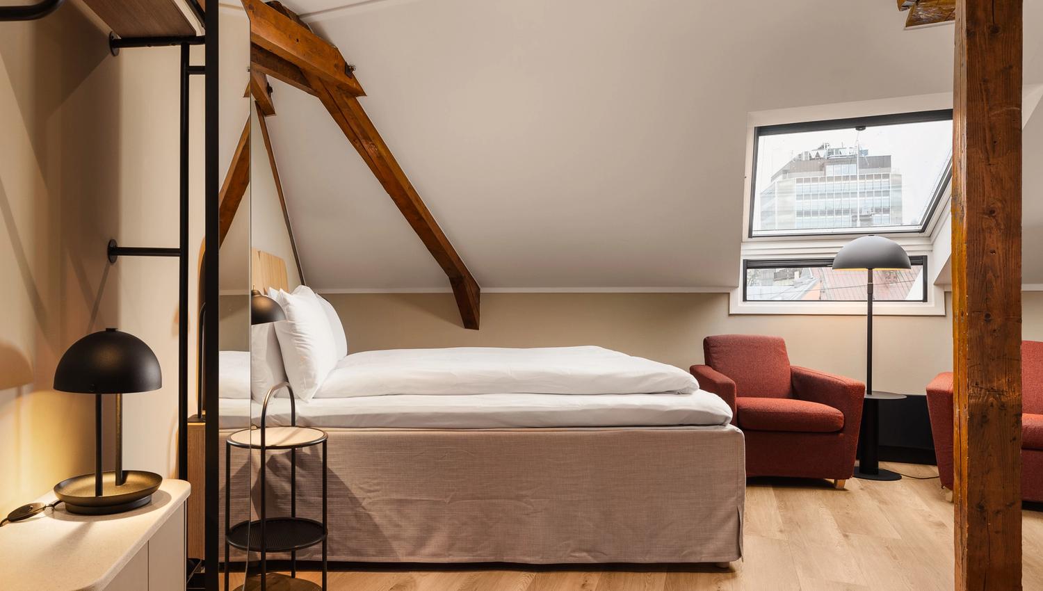 A hotel room interior with a double bed, a window, brown armchairs and a brown ceiling beam running along the curved ceiling