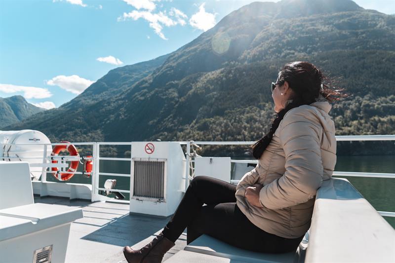 Passasjer sit på utedekket i solskinn under fjordcruise på Hardangerfjorden med grøne fjellsider rundt.