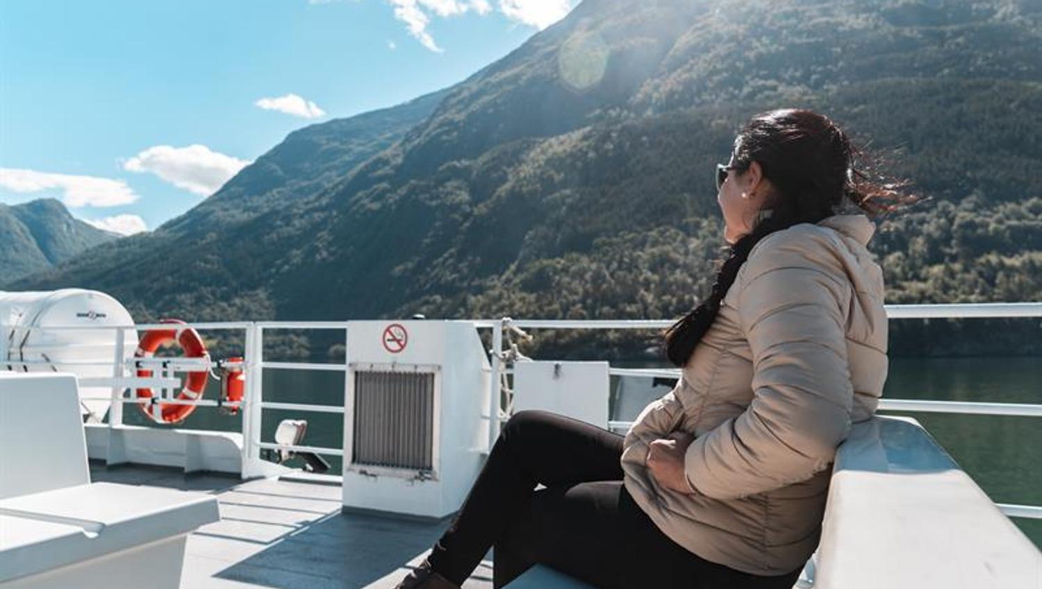 Passasjer sit på utedekket i solskinn under fjordcruise på Hardangerfjorden med grøne fjellsider rundt.