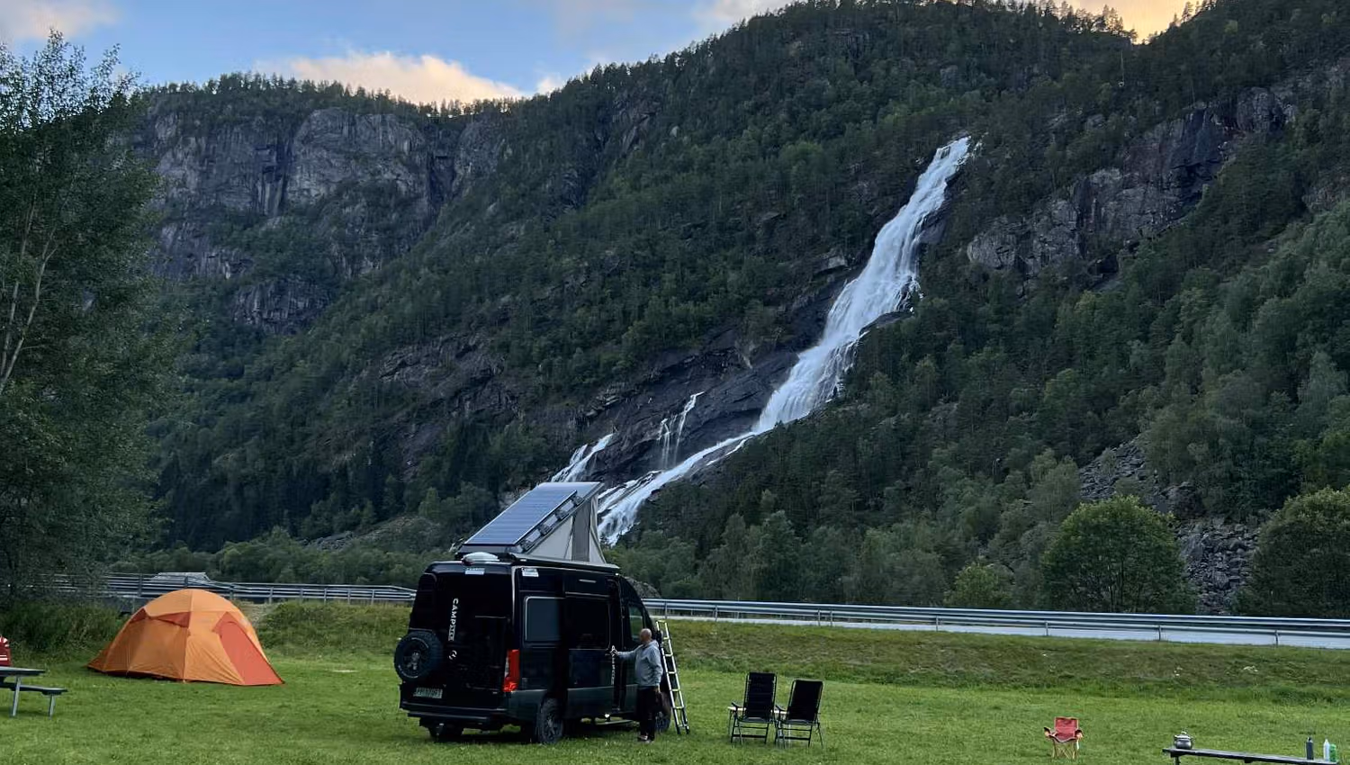 Wohnmobil und Zelte auf dem Hildal Camping mit einem beeindruckenden Wasserfall im Hintergrund.