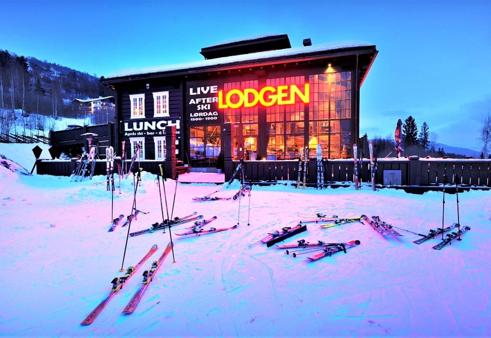 Snötäckt skidområde i skymning med lodgebyggnad med upplysta fönster, neonskylt och skidor i förgrunden.