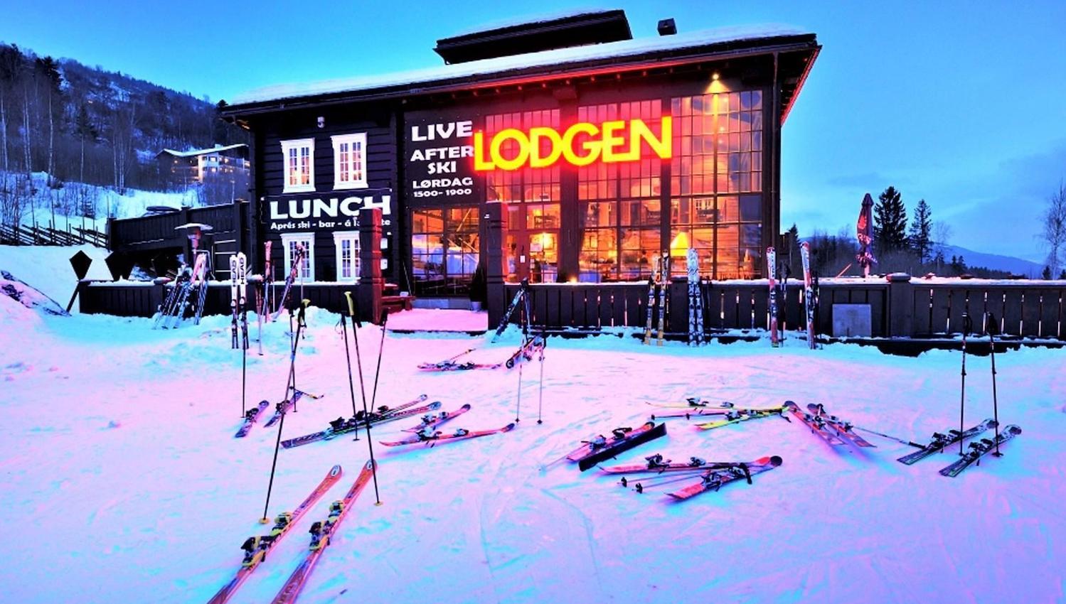 Snötäckt skidområde i skymning med lodgebyggnad med upplysta fönster, neonskylt och skidor i förgrunden.