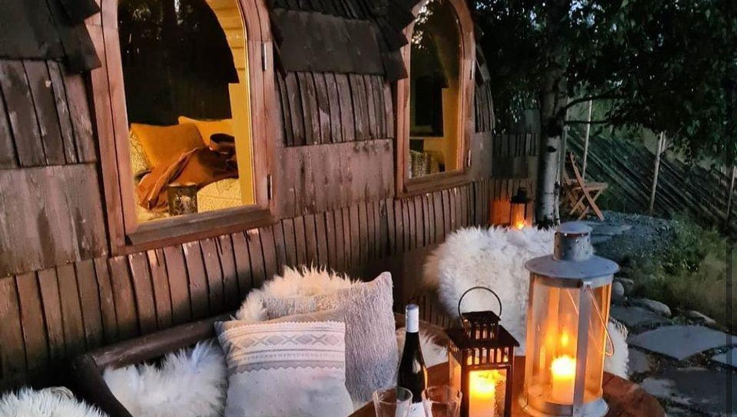 A cozy outdoor seating area with wooden benches, cushions, furs, and lit lanterns.