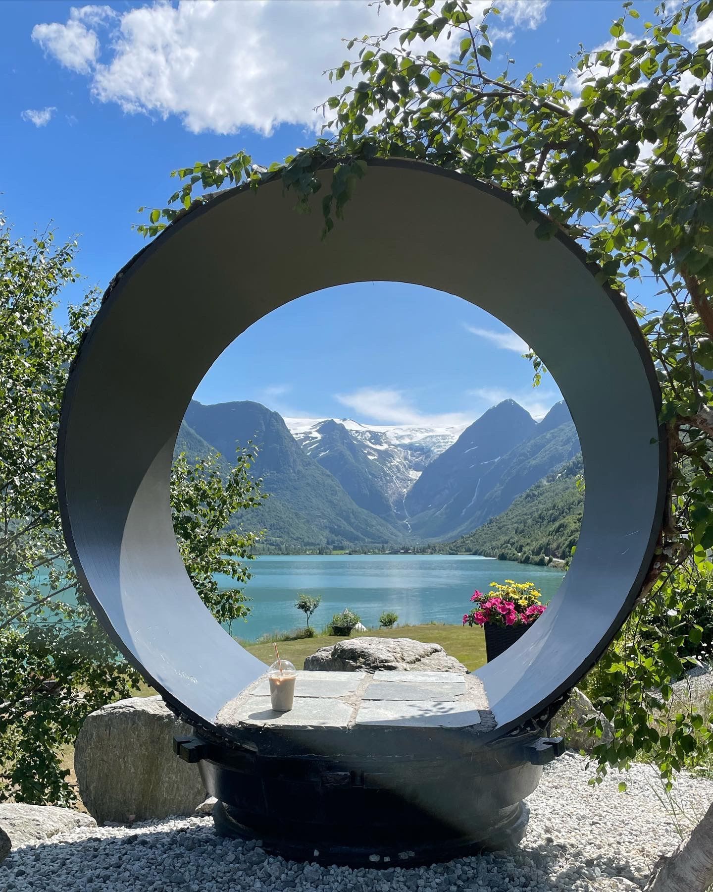 Large circular sculpture frames a turquoise lake and snowcapped mountains, with trees, rocks, and a drink in front.