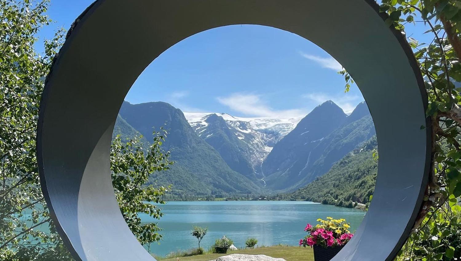 Large circular sculpture frames a turquoise lake and snowcapped mountains, with trees, rocks, and a drink in front.