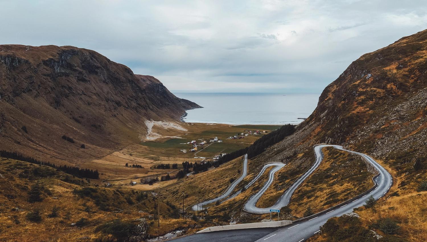 Svingete veg ned ein fjelldal mot fjord og hav, med ein bil i forgrunnen og skyer over fjella..