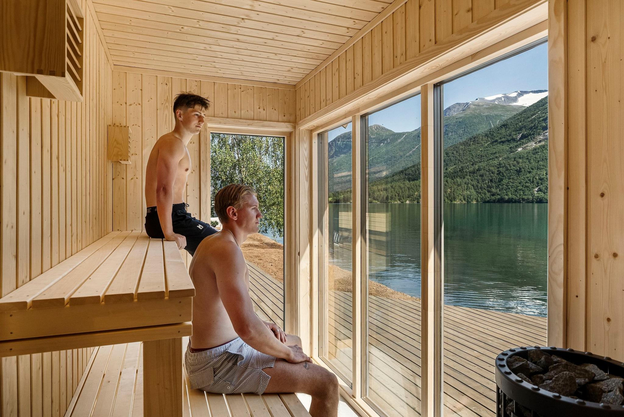 To menn sit i ei trebadstue med store glasdører mot Oppstrynsvatnet, stille vatn og grøne fjell i sollys..