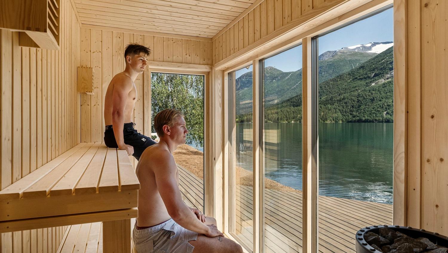 Two shirtless men sit on wooden benches inside a sauna, looking through large windows at a lake and mountains..