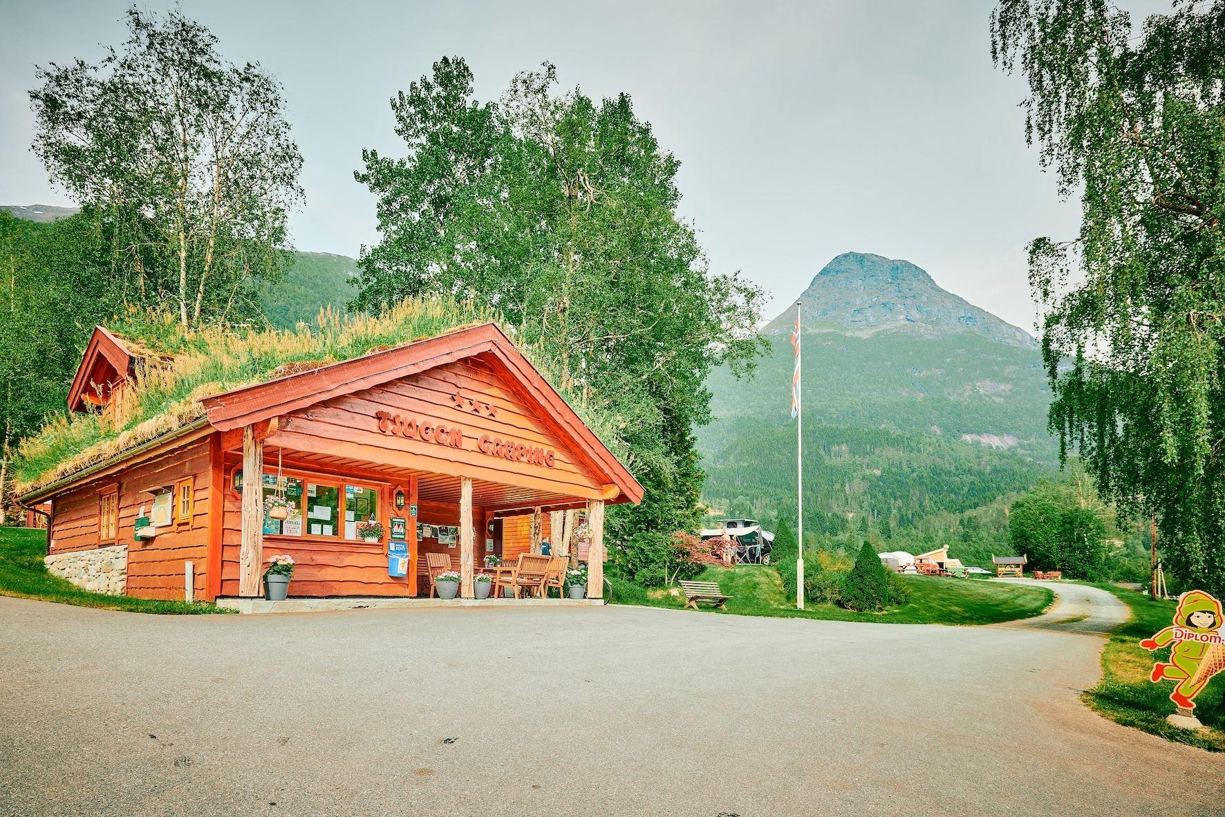 Red wooden campsite lodge with grass roof by a paved road, trees and a mountain landscape in the background..