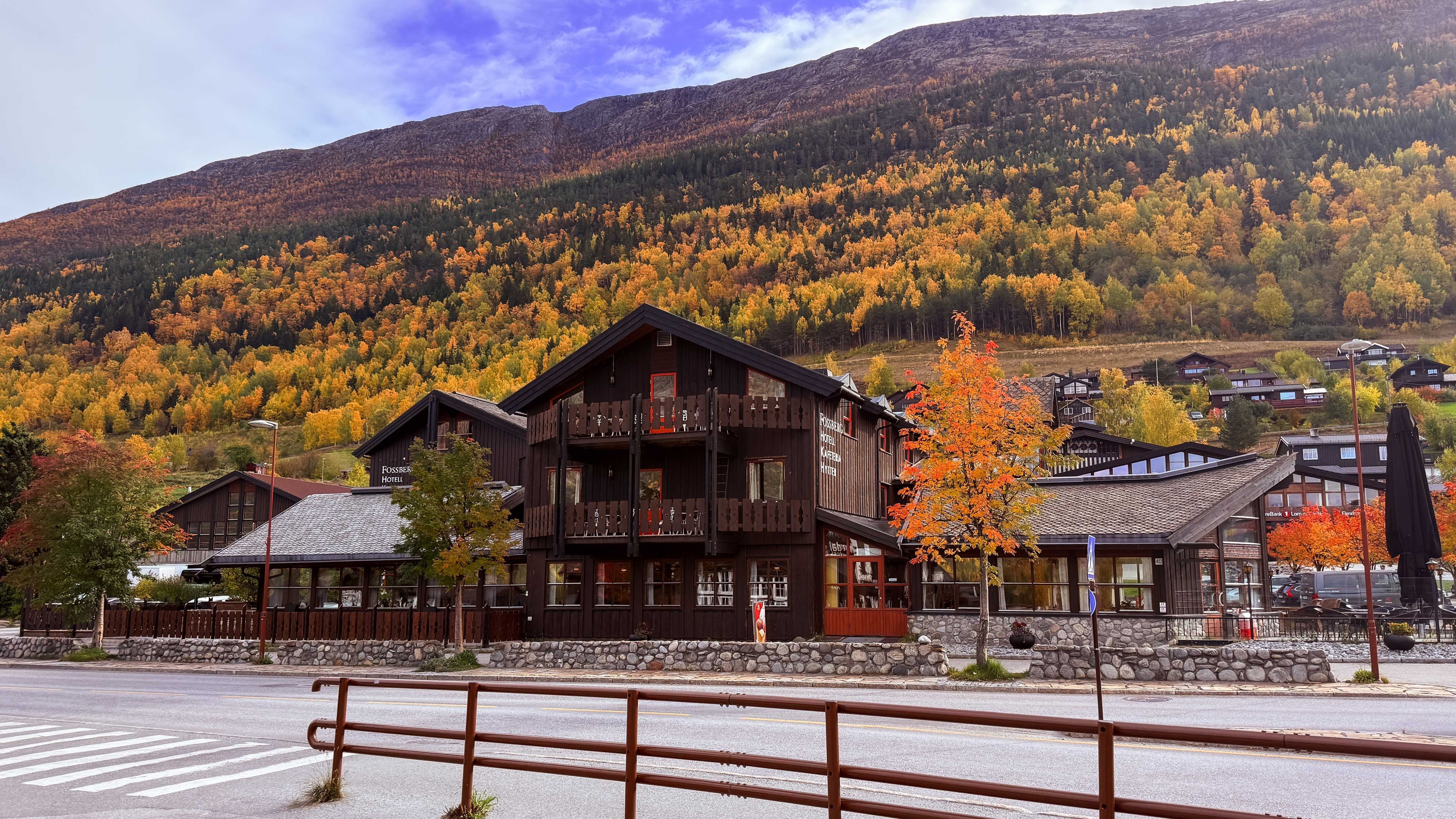 Dark wooden hotel buildings by a road, with orange autumn trees and a forested mountain slope under a cloudy sky..