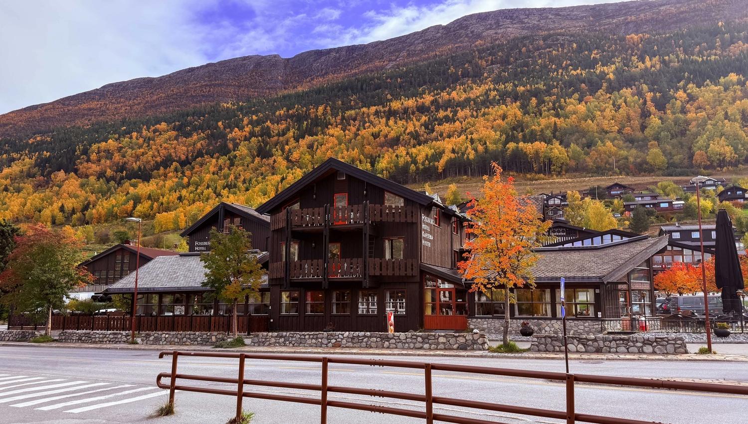 Dark wooden hotel buildings by a road, with orange autumn trees and a forested mountain slope under a cloudy sky..