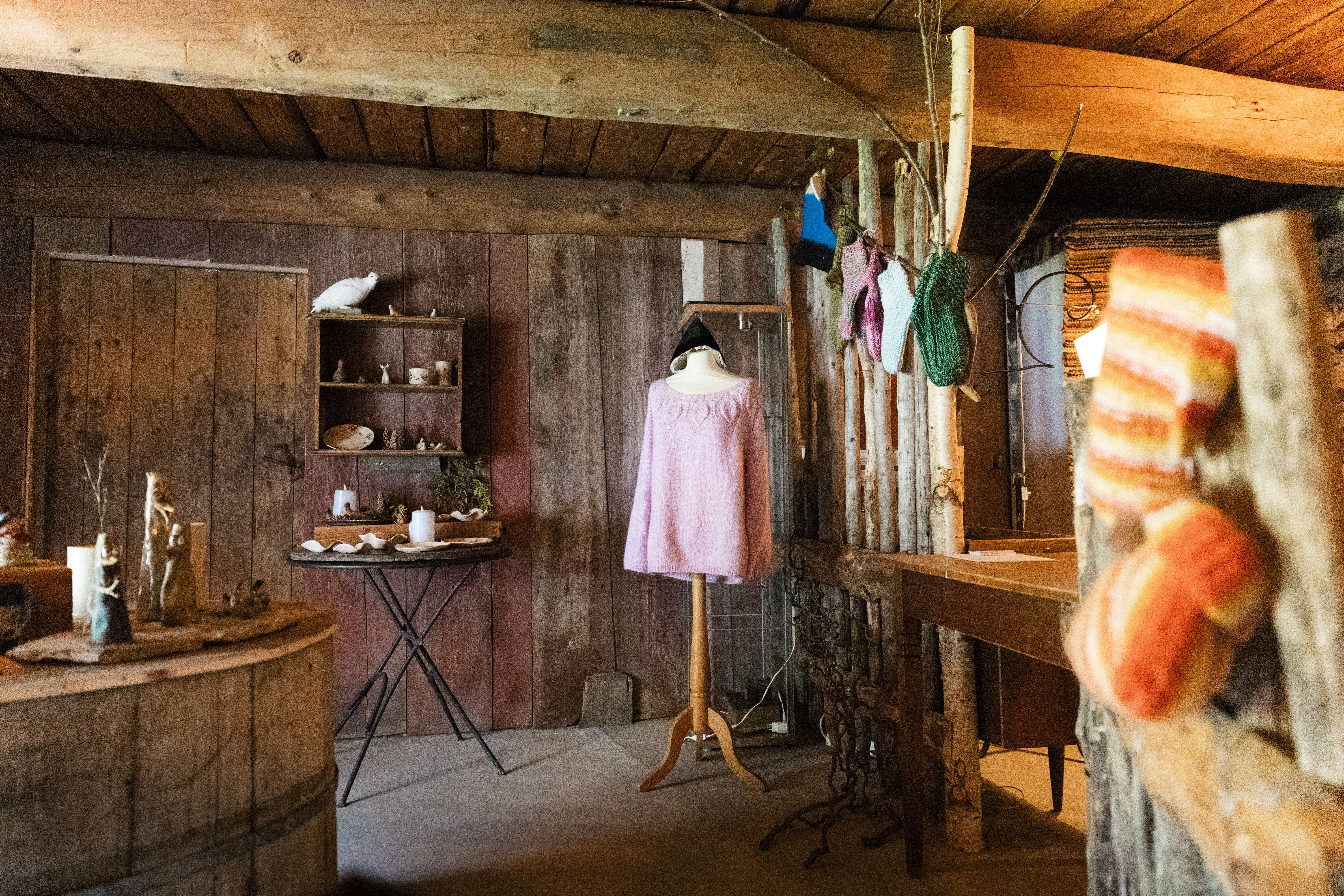 Warm-lit rustic wooden room with a mannequin in a pink sweater, shelves of small items, and hanging yarn.
