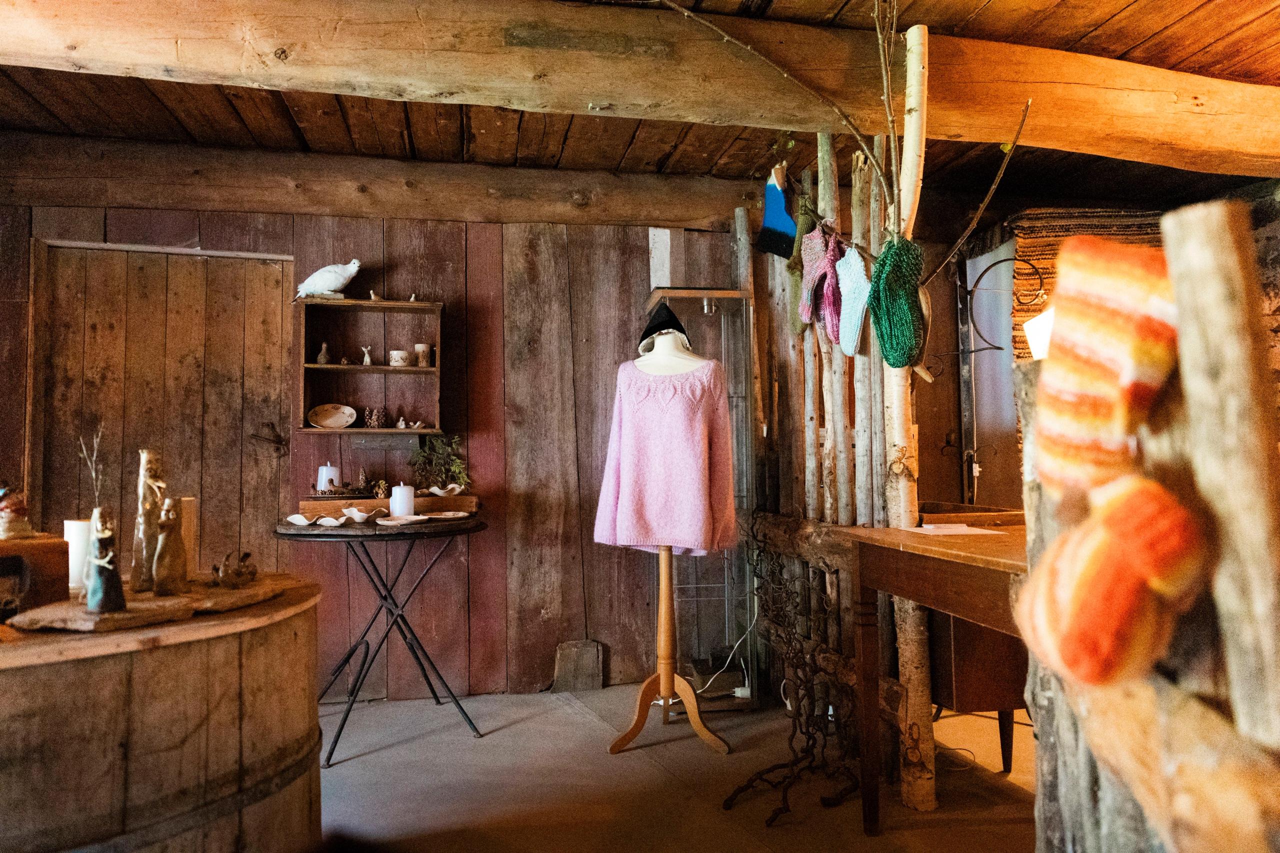 Warm-lit rustic wooden room with a mannequin in a pink sweater, shelves of small items, and hanging yarn.