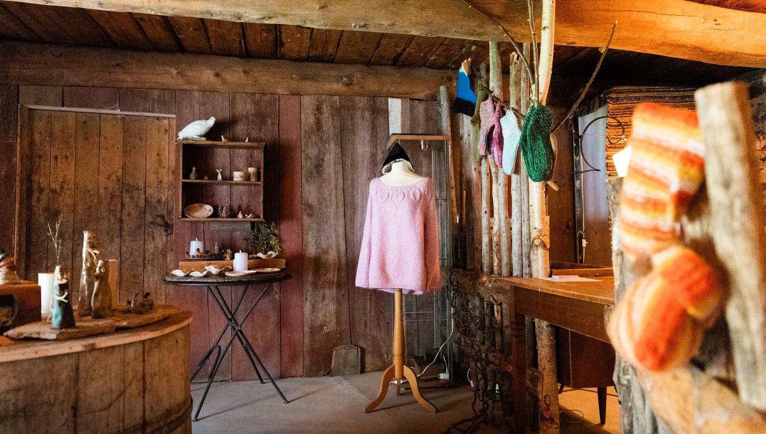 Warm-lit rustic wooden room with a mannequin in a pink sweater, shelves of small items, and hanging yarn.
