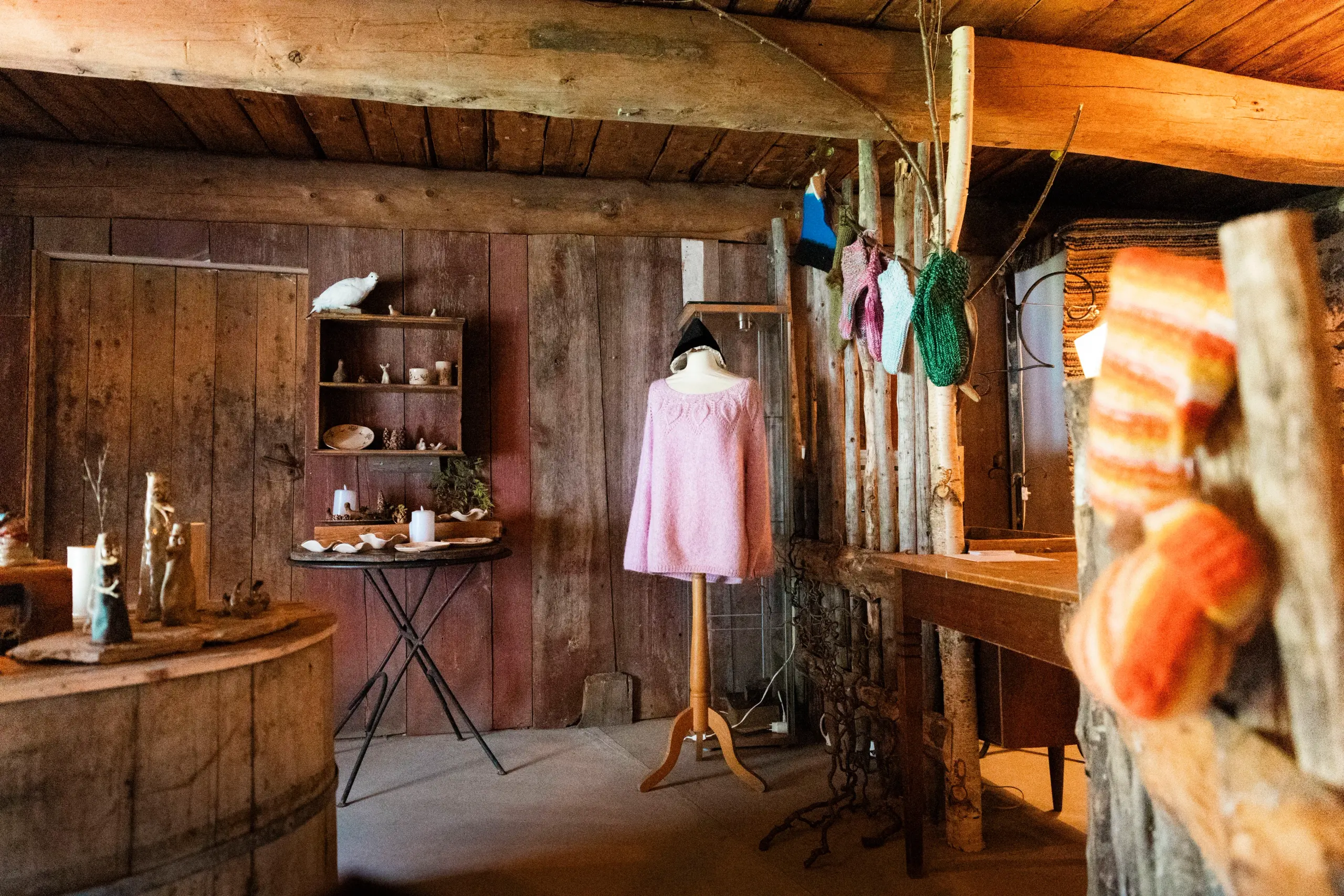 Warm-lit rustic wooden room with a mannequin in a pink sweater, shelves of small items, and hanging yarn.