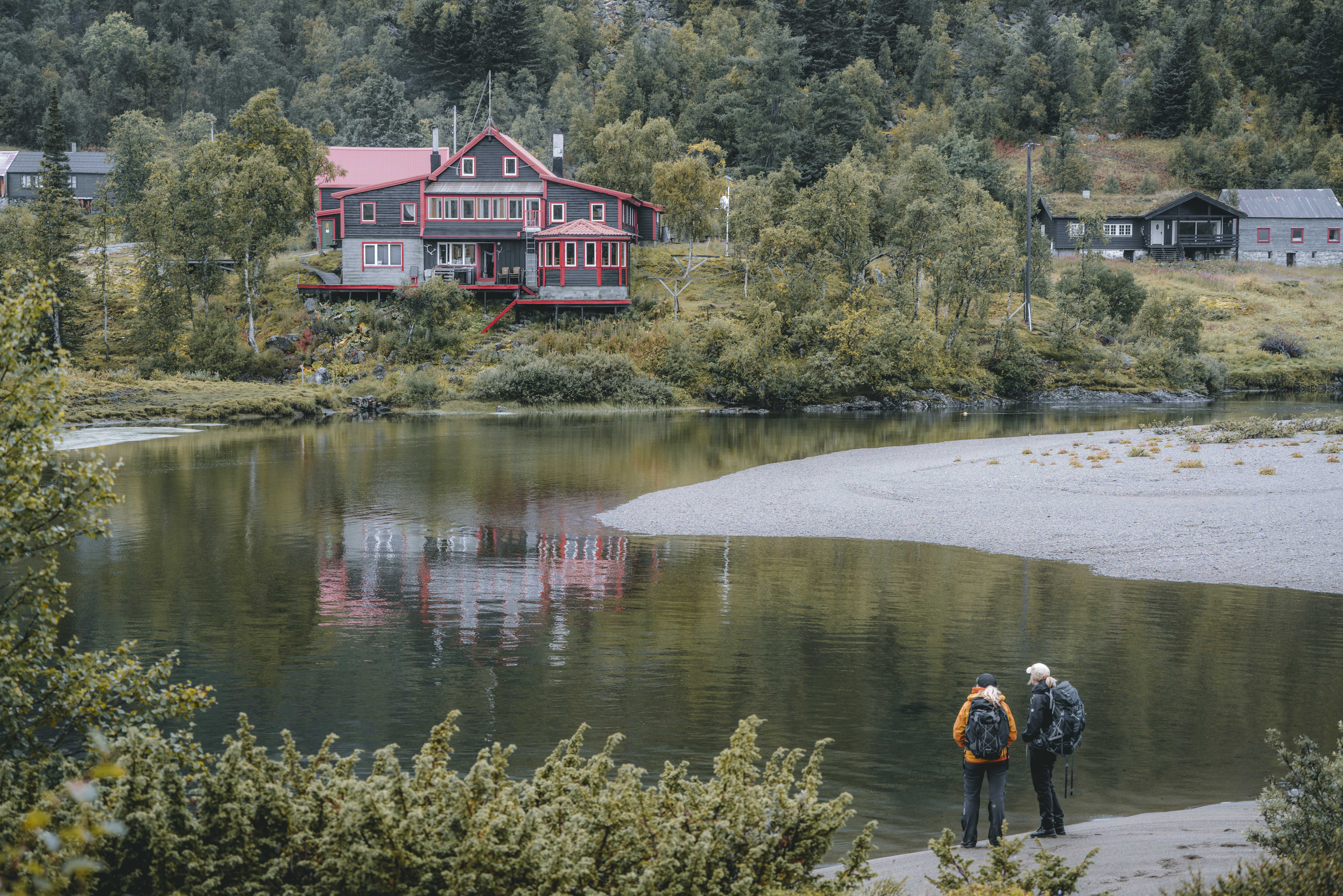 Zwei Frauen stehen am Fluss und blicken auf die Vivelid-Berghütte, die auf der anderen Flussseite liegt.