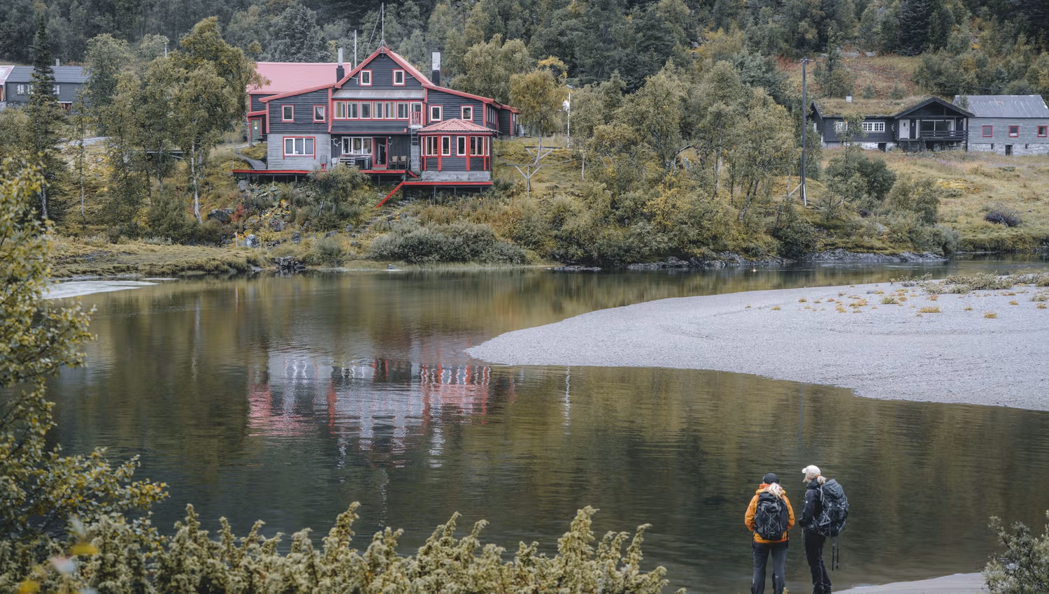 Zwei Frauen stehen am Fluss und blicken auf die Vivelid-Berghütte, die auf der anderen Flussseite liegt.